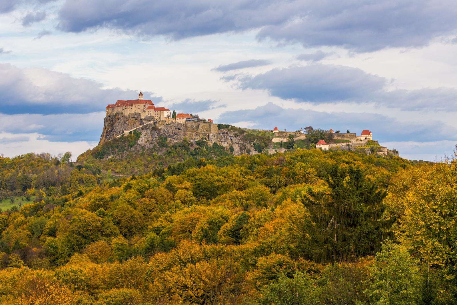 Ein herbstlicher Wald im Vordergrund. Dahinter ergebt sich ein Felsen mit einer Burg on top. Der Himmel ist bewölkt.