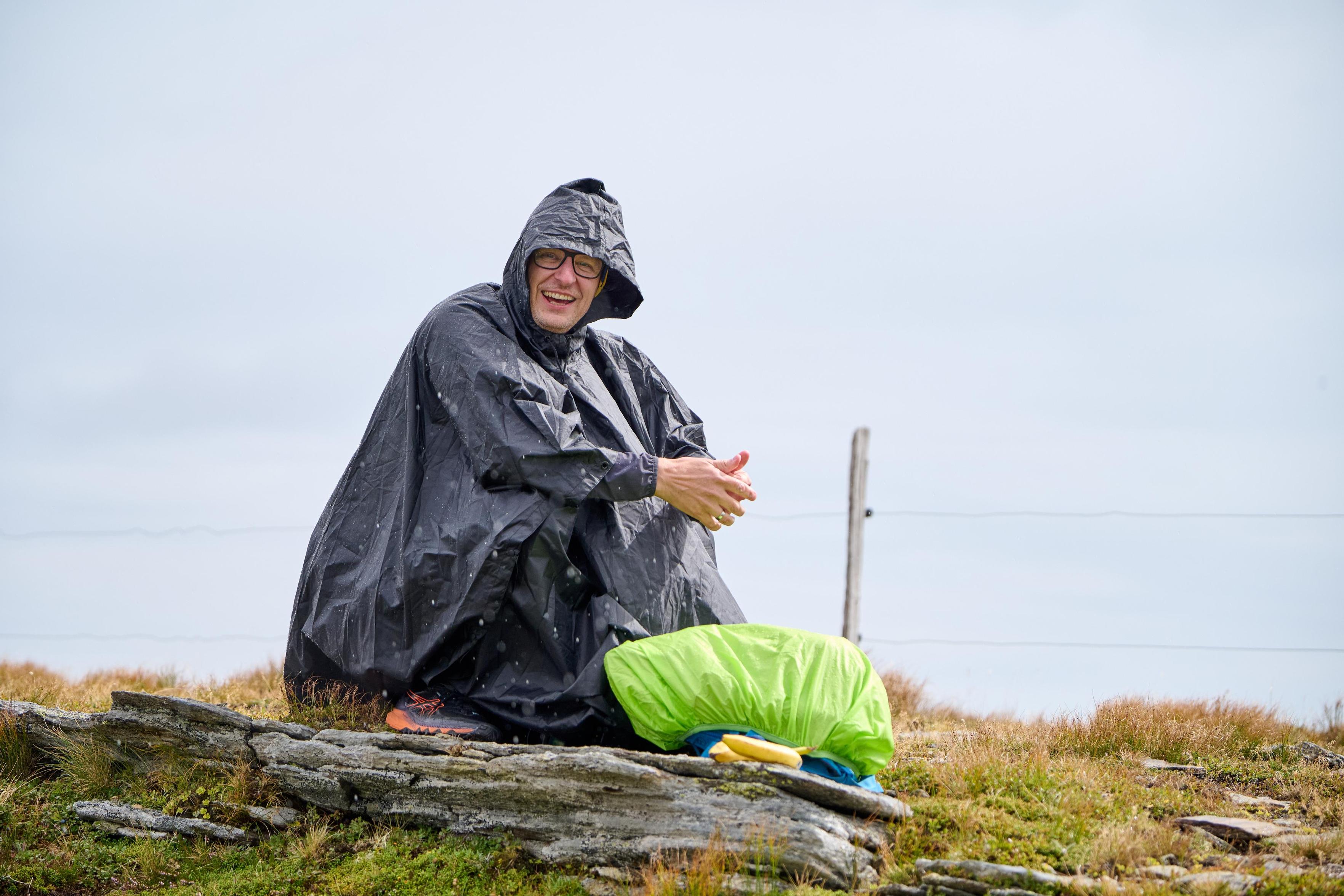 Ein Mann hockt in einen Regenponcho gehüllt hinter einem Rucksack, der ebenfalls im Regenschutz eingehüllt ist, am Boden. Er zeigt Daumen hoch, obwohl es offensichtlich regnet. Es ist eine Berggegend, dahinter ist ein Weidezaun zu erkennen, der Himmel ist bewölkt.