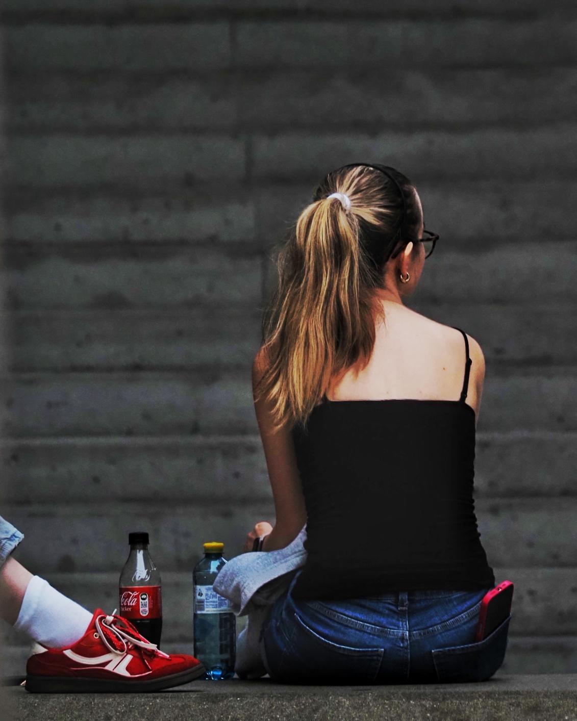 Eine Frau sitzt mit dem Rücken zur Kamera, links davon stehen eine Wasser- und eine Colaflasche. Von links sieht man noch rote Schuhe, die wohl zu einer weiteren Person gehören.