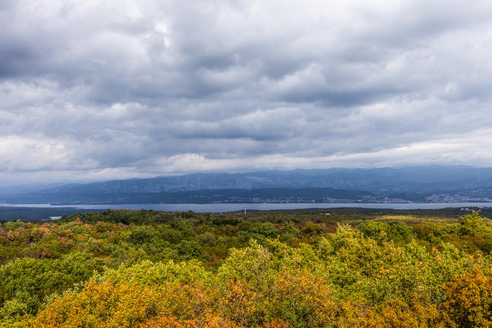 Deutsch: Das Bild zeigt eine weite, malerische Landschaft mit einer Mischung aus üppigem Grün und herbstlichen Farbtönen im Vordergrund. Im Hintergrund erstreckt sich ein großer See oder eine Bucht, umgeben von bergigen Erhebungen, die in bläulichen und grauen Tönen schimmern. Der Himmel ist bewölkt, mit dichten, dunklen Wolken, die eine dramatische Stimmung erzeugen. Die Szene wirkt friedlich, aber auch leicht melancholisch durch das Spiel von Licht und Schatten.

English: The image depicts a vast, picturesque landscape with a mix of lush greenery and autumnal hues in the foreground. In the background, a large lake or bay stretches out, surrounded by mountainous ridges that shimmer in bluish and gray tones. The sky is overcast, with dense, dark clouds creating a dramatic atmosphere. The scene feels serene yet slightly melancholic due to the interplay of light and shadow.